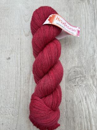 Ein Strang handgefärbter Wolle in einem warmen, tiefen Rotton liegt auf hellem Holz. Der Strang ist gedreht, die Melierung der Faser ist sichtbar, die Herzfasern-Banderole ist umgelegt.